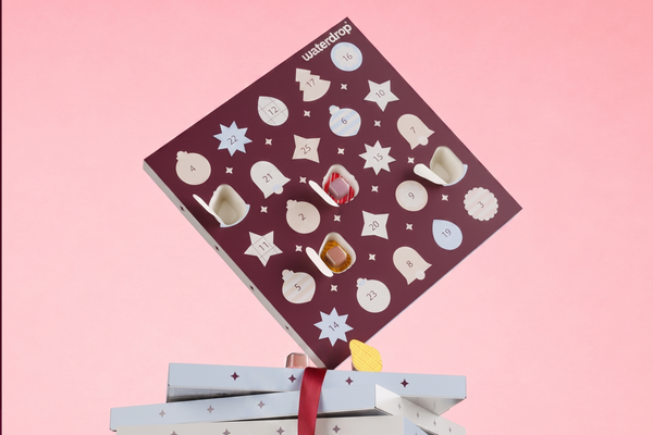 Advent kalendar sa kružnim prozorima i malim predmetima iznad poklona na ružičastoj pozadini.